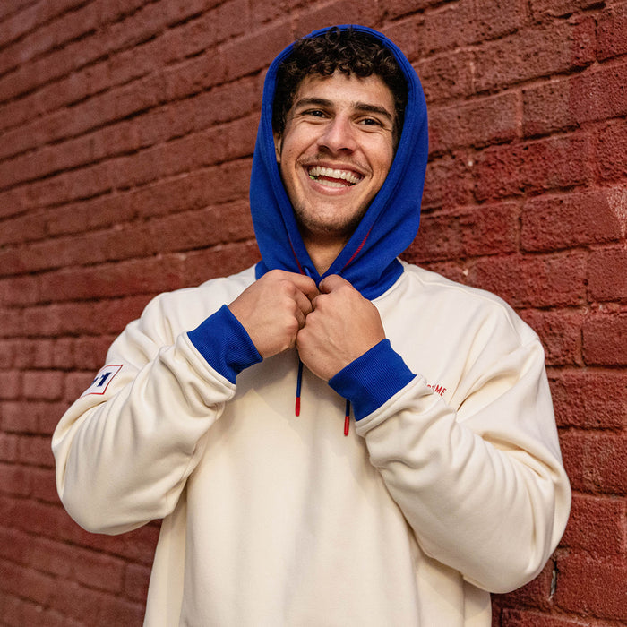 Smiling man pulling up the royal blue hood of a cream stadium hoodie against a brick wall