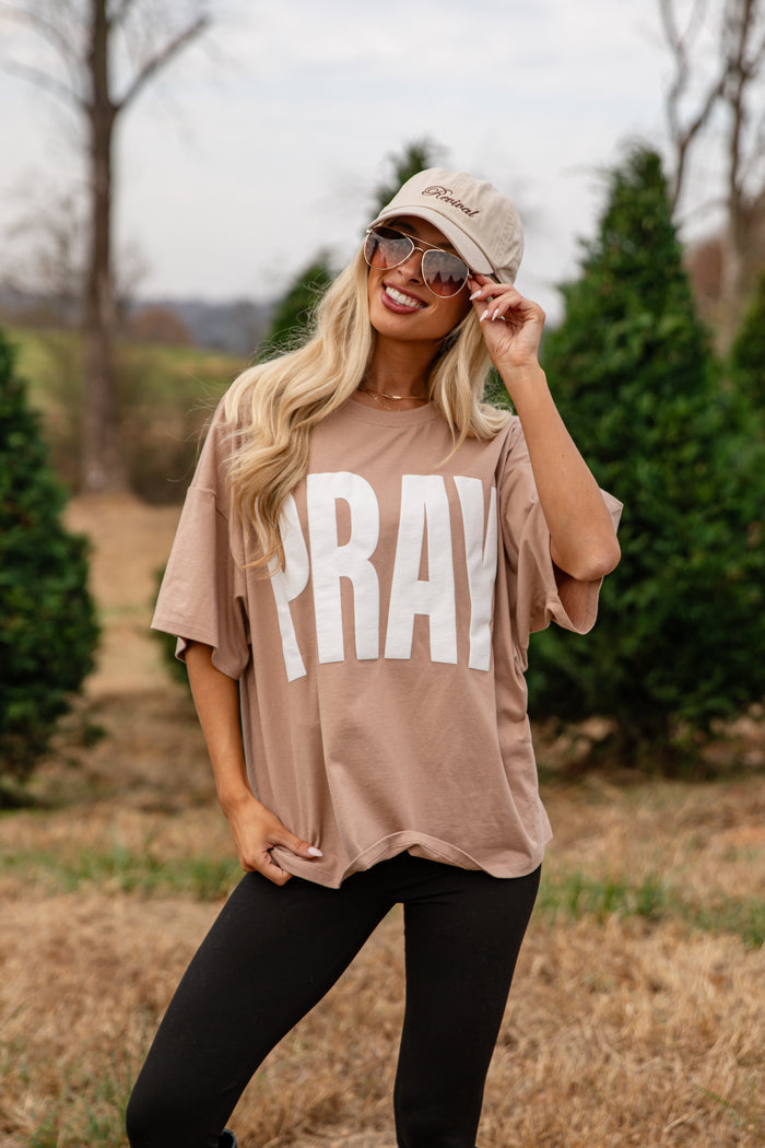 Woman wearing a beige 'PRAY' shirt outdoors with trees in the background