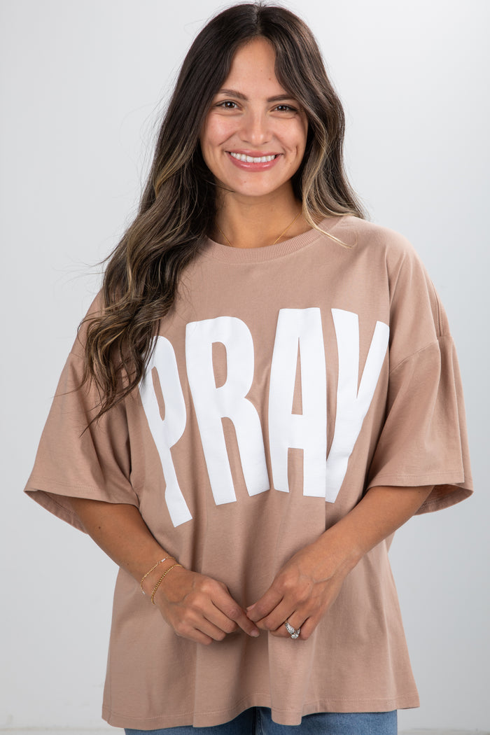 Woman wearing a beige t-shirt with 'PRAY' printed on it against a white background