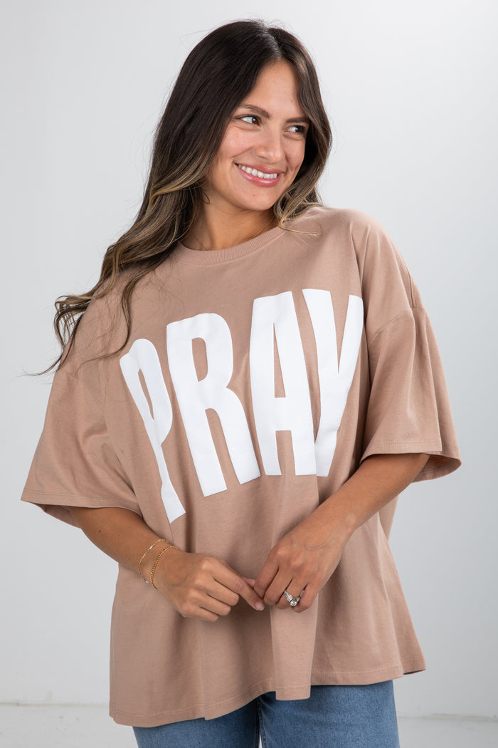 Woman wearing a beige t-shirt with 'PRAY' printed in white on a plain background